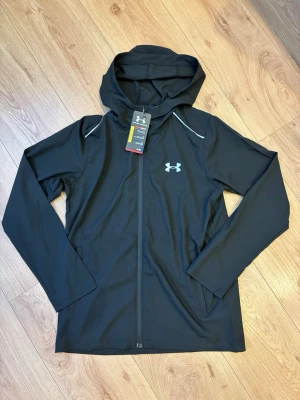Under Armour jacka  - Sportig svart löparjacka från Under Armour med huva och hel dragkedja framtill. Jackan har reflekterande detaljer vid axlarna och logga på bröstet. Tillverkad i lätt och funktionellt material som andas, perfekt för löpning och träning utomhus.