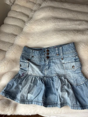 Blå volang jeanskjol med fickor - Säljer en kort jeanskjol i ljusblå denim med volang nedtill och flera snygga fickor fram. Kjolen har dekorativa knappar i midjan och coola sömdetaljer. Perfekt för dig som gillar en avslappnad men trendig stil.