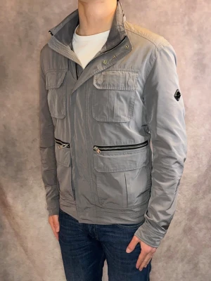 Grå J-Lindeberg Fieldjacket Vår/Höst - Stl M - Riktigt snygg Fieldjacket från J-Lindeberg i färgen grå! | Mycket bra skick: 9/10, inga defekter | Stl: M, modellen är 177cm och väger 75kg, passar riktigt bra 🙌  | Hör av dig vid frågor // Galgen 🫡