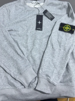 Grå sweatshirt från Stone Island - Snygg grå sweatshirt från Stone Island med klassisk rund hals och ribbade muddar. Tröjan har den ikoniska Stone Island-loggan på vänster ärm och är tillverkad i mjuk bomullsblandning. Perfekt för en avslappnad och trendig stil.