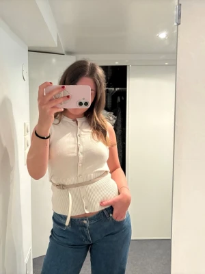 Beige stickad väst med knappar - Snygg beige stickad väst med knappar framtill och ribbad midja. Västen har en dragsko i midjan för justerbar passform och är ärmlös. Perfekt att styla över en t-shirt eller skjorta för en trendig look.