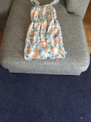 Blommig sommarklänning med volang - Söt klänning med blommigt mönster i orange, grönt och vitt på ljusblå botten. Klänningen har smala axelband, volangdetaljer och smock i midjan för en bekväm passform. Perfekt för varma dagar och ger en fräsch och ungdomlig look.