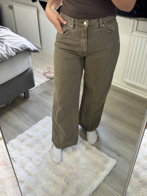 Olivgröna wide leg jeans - Säljer ett par olivgröna jeans från Gina med hög midja och raka, vida ben. Jeansen har klassiska fem fickor och stängs med dragkedja och knapp. Materialet är kraftigt denim som ger en snygg och avslappnad look.