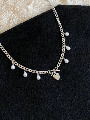 Silverfärgat halsband med pärldroppar - Snyggt silverfärgat halsband rostfritt stål med små vita pärldroppar och ett hjärthänge med rosett. Kedjan har en chunky design och pärlorna hänger jämnt längs hela halsbandet. Perfekt accessoar för att lyfta din outfit.