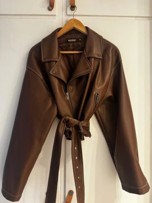 Brun skinnjacka med bälte från Bianca - Snygg brun skinnjacka från Bianca med bred krage, asymmetrisk dragkedja och bälte i midjan. Jackan har en cool bikerstil med dragkedjedetaljer och markerade sömmar. Perfekt för dig som vill ha en trendig och edgy look.