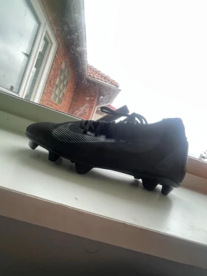 Svarta Nike Mercurial academy fotbollsskor - Säljer ett par svarta Nike Mercurial fotbollsskor med snörning och dobbar för spel på gräs. Skorna har en modern, strömlinjeformad design med diskreta detaljer och Nike-logga på sidan. Tillverkade i syntetmaterial för bra grepp och känsla på planen.