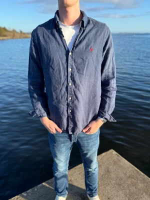Blå linneskjorta från Ralph Lauren - Snygg blå skjorta från Ralph Lauren i linne med klassisk krage och knappar framtill. Liten röd logga broderad på bröstet. Långärmad modell med avslappnad passform, perfekt till jeans eller chinos. Storlek L men passar M också! Väldigt bra skick och vårt pris 449kr men kan gå ner vid snabb och smidig affär💪 // Studio II Resell 💙