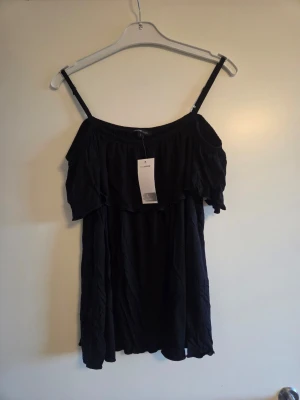 Svart axelbandslös volangtopp, nyskick, stl L - Snygg svart linnetopp med tunna axelband och volangdetalj över bysten. Toppen är axelbandslös i stilen och har en loose passform som ger en avslappnad vibe. Perfekt för sommardagar eller när du vill känna dig extra chill.
