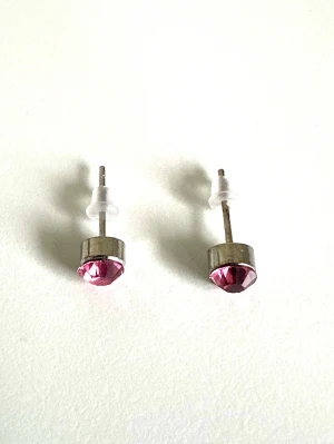 Silvriga örhängen med rosa sten, HELT NYA - Snygga små örhängen i silverfärgad metall med rund, rosa glittrande sten framtill. Klassisk och enkel design med plastpluppar som lås. Perfekta för dig som gillar diskreta men färgglada accessoarer. Säljes helt NYA i förpackning pga dubblett. 