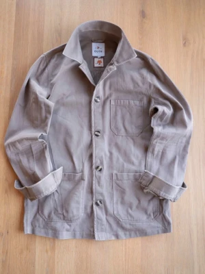 Beige overshirt från OLOF - Stilren beige overshirt från OLOF med klassisk krage och tre framfickor. Tillverkad i mjuk bomull och har knäppning framtill. Perfekt lager-på-lager-plagg för en avslappnad look.