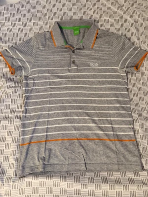 Grå randig t-shirt från BOSS - Snygg grå t-shirt från BOSS med vita horisontella ränder och orange detaljer vid krage och ärmslut. Klassisk pikékrage och knappslå framtill. Perfekt för en avslappnad men stilren look.