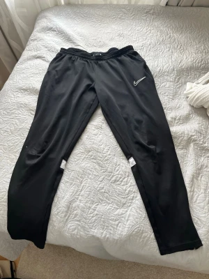 Svarta träningsbyxor från Nike - Säljer ett par svarta träningsbyxor från Nike med vit broderad logga på vänster lår. Byxorna har resår i midjan och smal passform med vita detaljer vid knäna. Perfekta för gymmet eller löprundan. De har en liten skada vid knät men annars väldigt bra skick 