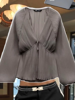 Plisserad grå blus från Zara - Trendig slutsåld blus från Zara med djup v-ringning och knytning framtill. Blusen har vida, långa ärmar och ett flowy, avslappnat fall. Perfekt för dig som gillar en modern och chic stil.