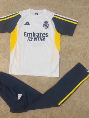 Real Madrid träningsset Adidas - Snyggt träningsset från Real Madrid och Adidas. Tröjan är vit med gula och mörkblå detaljer, klubbmärke och Emirates Fly Better-tryck. Byxorna är mörkblå med gula ränder längs sidorna och Adidas-logga. Perfekt för fotbollsträning eller chill.