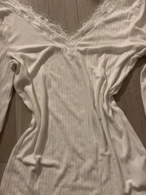 Vit spetstopp - Säljer en vit långärmad topp med ribbad struktur och v-ringning dekorerad med spets. Toppen har en feminin och smickrande passform, perfekt för att lyfta vilken outfit som helst. Materialet känns mjukt och stretchigt. 