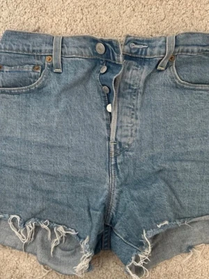 Levi's Premium blå jeansshorts - Säljer ett par klassiska blå jeansshorts från Levi's Premium med hög midja, råa kanter och knappgylf. Shortsen har fem fickor och är tillverkade i slitstark bomull. Perfekta för sommardagar och ger en avslappnad look.