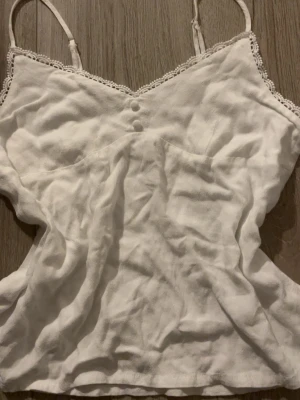 Vitt linne  - Söt vit linnetopp med tunna axelband och spetsdetaljer längs urringningen. Toppen har två små dekorativa knappar framtill och är tillverkad i ett lätt material, perfekt för varma dagar.