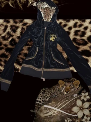 Svart hoodie med leopardmönster/roberto cavalli - Snygg svart hoodie från roberto  med guldfärgade detaljer och dragkedja framtill. Huvan är fodrad med leopardmönstrat tyg och bröstet har en guldfärgad logga. Två fickor fram och ribbade muddar vid ärmslut och nederkant. Perfekt för dig som gillar djurmönster!