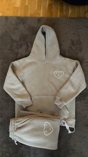 Carsicko Tracksuit - Mysigt Grå Mjukis-set med hoodie och matchande byxor. Båda plaggen har en vit broderad hjärtdetalj med ansikte på bröstet och benet. Hoodien har känguruficka och byxorna har resår i midjan med dragsko. Perfekt för chill dagar hemma.