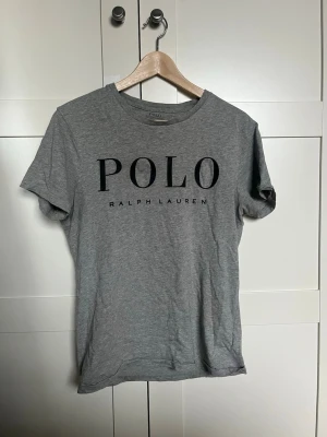 Grå t-shirt från Polo Ralph Lauren - Snygg grå t-shirt från Polo Ralph Lauren med stor svart logga framtill. Klassisk rund halsringning och korta ärmar. Tillverkad i mjuk bomull för en skön känsla och enkel stil.