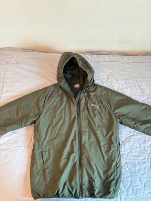 Olivgrön vindjacka från Puma S - Snygg olivgrön vindjacka från Puma med huva och dragkedja framtill. Jackan har två sidofickor och elastisk nederkant. Tillverkad i lätt och tåligt material som skyddar mot vind. Perfekt för dig som vill ha en sportig och stilren look.