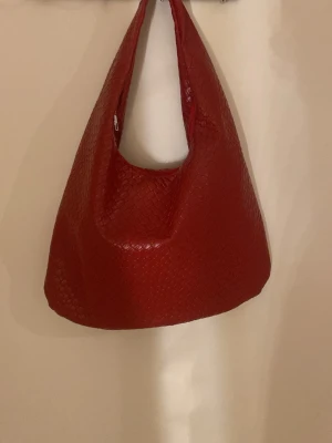 Röd axelväska i flätad look - Snygg röd handväska med axelrem och flätad struktur. Väskan har en mjuk, slouchy form och stängs med dragkedja. Materialet ser ut som konstläder med en cool textur som ger extra stilpoäng.