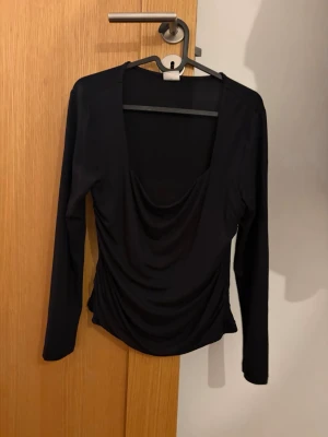 Svart långärmad topp med drapering - Snygg svart långärmad topp med djup urringning och draperad front. Toppen har en figurnära passform och är tillverkad i ett mjukt, stretchigt material som ger en smickrande siluett. Perfekt att styla med jeans eller kjol för en trendig look.