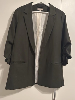 Oliv  oversized kavaj med fickor - Svart oversized kavaj med klassisk krage och två raka fickor framtill. Kavajen har vit slät innerfoder och uppvikta ärmar för en avslappnad look. Perfekt för dig som gillar en trendig och chill stil.