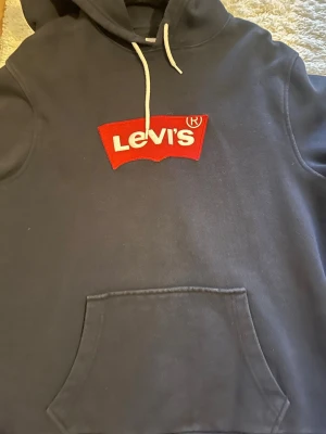Mörkblå Levi's hoodie med logga - Snygg mörkblå hoodie från Levi's med stor röd och vit logga på bröstet. Klassisk känguruficka framtill och vit dragsko i huvan. Tillverkad i mjukt bomullsmaterial som är perfekt för chill dagar. Enkel och stilren design som funkar till det mesta.