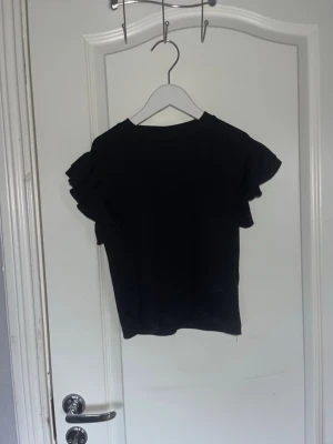 Svart topp med volangärmar - Snygg svart topp från Gina Tricot med korta volangärmar och rund hals.