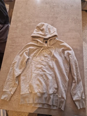 Grå hoodie från GANT med logga - Snygg grå hoodie från GANT med broderad logga på bröstet och klassisk känguruficka. Hoodien har dragsko i huvan och ribbade muddar vid ärmar och nederkant. Perfekt för en chill och avslappnad stil.