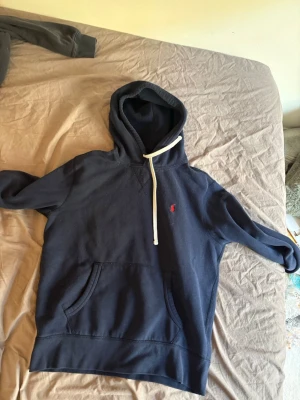 Mörkblå hoodie från Polo Ralph Lauren - Snygg mörkblå hoodie från Polo Ralph Lauren med klassisk röd logga broderad på bröstet. Hoodien har vit dragsko i huvan och en stor magficka framtill. Tillverkad i mjukt bomullsmaterial som är perfekt för chill dagar.