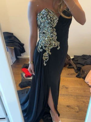 Svart långklänning med paljetter - Säljer en svart axelbandslös långklänning med glittriga paljettdetaljer i guld och silver över bysten och ena sidan. Klänningen har en snygg slits framtill och figurnära passform som ger en elegant siluett. Perfekt för bal eller festkvällar.