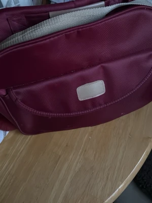 Vinröd ryggsäck med beige detaljer - Snygg vinröd ryggsäck med beige handtag och detaljer. Tillverkad i slitstarkt tygmaterial och har en enkel, stilren design med en liten beige patch framtill. Perfekt för dig som vill ha en praktisk och trendig väska till skolan eller stan.