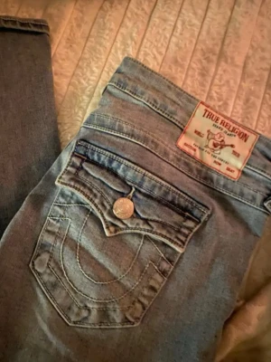 True Religion ljusblå jeans - Säljer ett par ikoniska ljusblå jeans från True Religion med deras klassiska broderade fickor och logopatch bak. Jeansen har raka ben och snygga detaljer på bakfickorna. Perfekta för en avslappnad och trendig look.