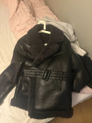 Svart skinnjacka med fuskpäls - Snygg svart skinnjacka med bred krage och detaljer i mörk fuskpäls. Jackan har bälte i midjan och pälsdetaljer vid ärmslut och nederkant. Perfekt för dig som vill ha en trendig och varm look i vinter.