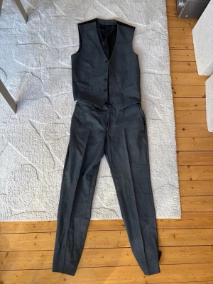 Mörkgrå kostymväst och byxa Dressmann - Snygg mörkgrå kostymväst och matchande byxa från Dressmann. Västen har knappar framtill och två fickor, byxorna har klassisk rak passform. Perfekt för dig som vill ha en stilren och enkel look. Materialet är polyester.