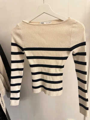 Randig stickad tröja från Zara - Säljer en krämvit stickad tröja från Zara med mörk blåa horisontella ränder. Tröjan har lång ärm, ribbad struktur och en något bredare båtringad hals. Materialet är mjukt och stretchigt, perfekt för lager på lager och en klassisk look.