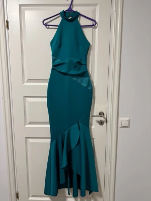 Turkos långklänning med halterneck - Snygg mörk turkos balklänning med halterneck och figurnära passform. Klänningen har volangdetaljer nedtill och en elegant, ärmlös design. Perfekt för speciella tillfällen där du vill sticka ut. Materialet känns slätt och har ett fint fall. 