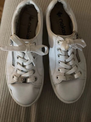 Vita sneakers från Rieker med dragkedja - Snygga vita sneakers från Rieker med klassisk låg profil, vita snören och dragkedja på sidan. Ovandelen är i vitt syntetmaterial och sulan är i gummi med brunt mönster undertill. Perfekta för en clean och fräsch look.