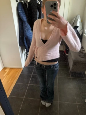 Ljusrosa omlottkofta med knytband - Supersöt ljusrosa stickad omlottkofta med knytband i midjan. Koftan har en croppad passform, långa ärmar och är gjord i ett mjukt, fluffigt material. Perfekt att styla över ett linne eller topp för en trendig look.