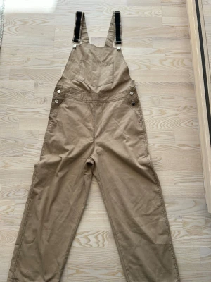 Beige ”snickarbrallor” från The Ragged Priest - Säljer ett par beige ”snickarbrallor” från The Ragged Priest i storlek S. Modellen har raka ben, justerbara svarta hängslen med metallspännen och fickor framtill. Tillverkad i ett slitstarkt bomullstyg som ger en cool och avslappnad look.