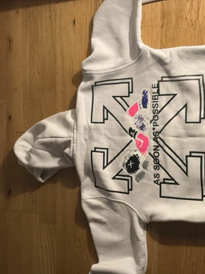 Vit Off-White hoodie med tryck - Vit hoodie från Off-White med stor grafisk pil-design och texten 'AS SOON AS POSSIBLE' på ryggen. Framsidan har Off-White-logga och rosa kryss. Hoodien har huva och magficka. Perfekt för dig som gillar streetwear och unika prints. Passar xs s och kanske m