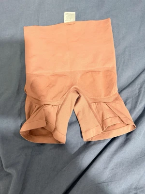 Nude shapewear shorts med hög midja - Säljer ett par nude shapewear shorts med hög midja och sömlös design. De är tillverkade i ett stretchigt material som formar och slätar ut siluetten. Perfekta under tighta kläder för en smidig look. Färgen är ljus beige vilket gör dem diskreta under de flesta plagg.