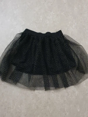 Kjol - Kort svart tyllkjol med elastisk midja och små guldprickar över hela kjolen. Kjolen har flera lager för extra volym och en glittrig look som sticker ut. Perfekt för dig som vill ha något extra till outfiten.