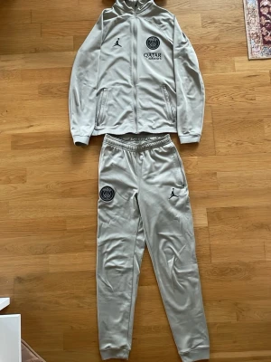 PSG träningsdress - Snygg grå träningsdress från PSG och Jordan med hel dragkedja på jackan och huva. Setet har PSG-logga och Jordan-logga på både jacka och byxa samt Qatar Airways-tryck. Tillverkad i mjukt, stretchigt material som passar perfekt till träning.
