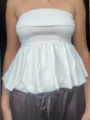 Vit axelbandslös tubtopp - Säljer en vit axelbandslös tubtopp med volang och ballongliknande form nedtill. Toppen har en bandeau-stil upptill och är perfekt för sommarens varma dagar. Materialet känns mjukt och stretchigt, troligen bomull eller syntet.