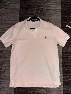 Vit pikétröja från Ralph Lauren - Relativt ny Klassisk vit pikétröja från Ralph Lauren med blå broderad logga på bröstet. Tröjan har krage, korta ärmar och knappar vid halsen. Perfekt för en clean och stilren look.