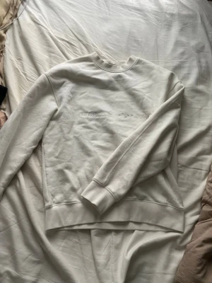 Vit sweatshirt med brodyr - Säljer en stilren vit sweatshirt med broderad text på bröstet och ryggen. Texten på ryggen listar stadsdelar i New York. Tröjan har rund halsringning, ribbade muddar och är tillverkad i mjukt bomullsmaterial. Perfekt för dig som gillar enkel men snygg streetwear.