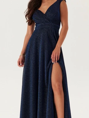 Marinblå glittrig långklänning - Marinblå långklänning med glittrigt tyg, v-ringad omlottdesign och breda axelband. Klänningen har en slits framtill och markerad midja som framhäver siluetten. Perfekt för bal, bröllop eller andra festliga tillfällen. Storlek M men notera att en skräddare kortat ner längden en aning, så den släpar lite baktill på någon som är ca 165cm. Använt ett fåtal gånger, lite slitage nertill i form av lösa trådar men inga större defekter. Kan självklart diskutera pris, hör av dig vid frågor! 